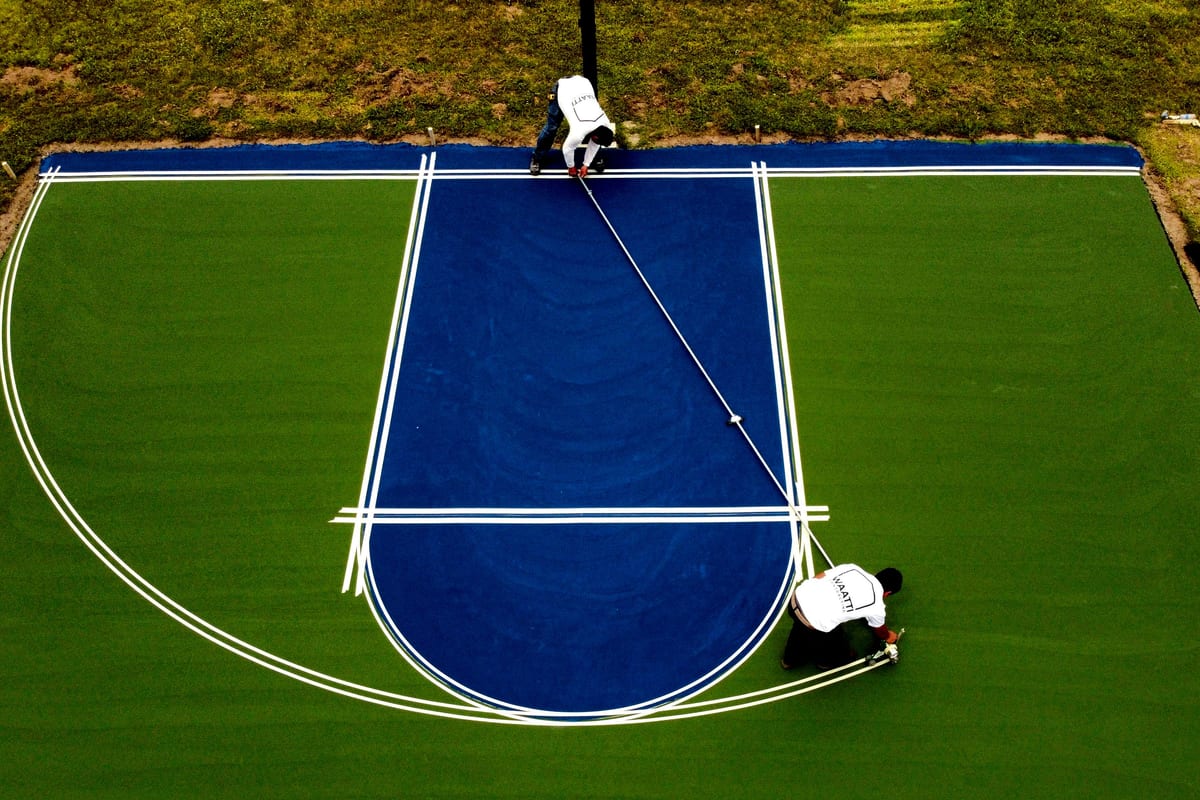 Drone view of two crew members taping and painting white key lines on a new blue and green basketball court — East Arcadia, NC