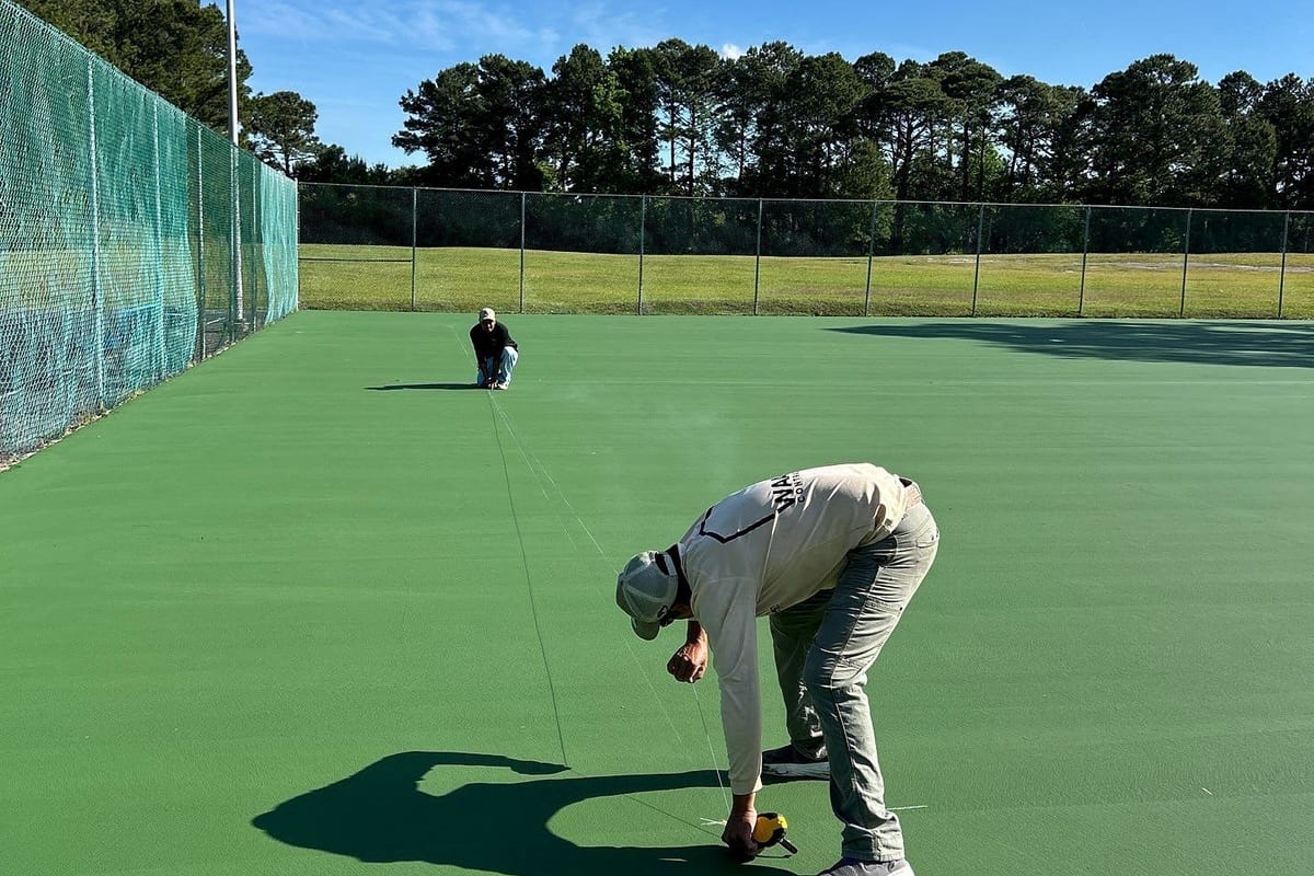 Crew member filling a crack on a green public tennis court before recoating, second crew member at the far fence — Castle Hayne, NC