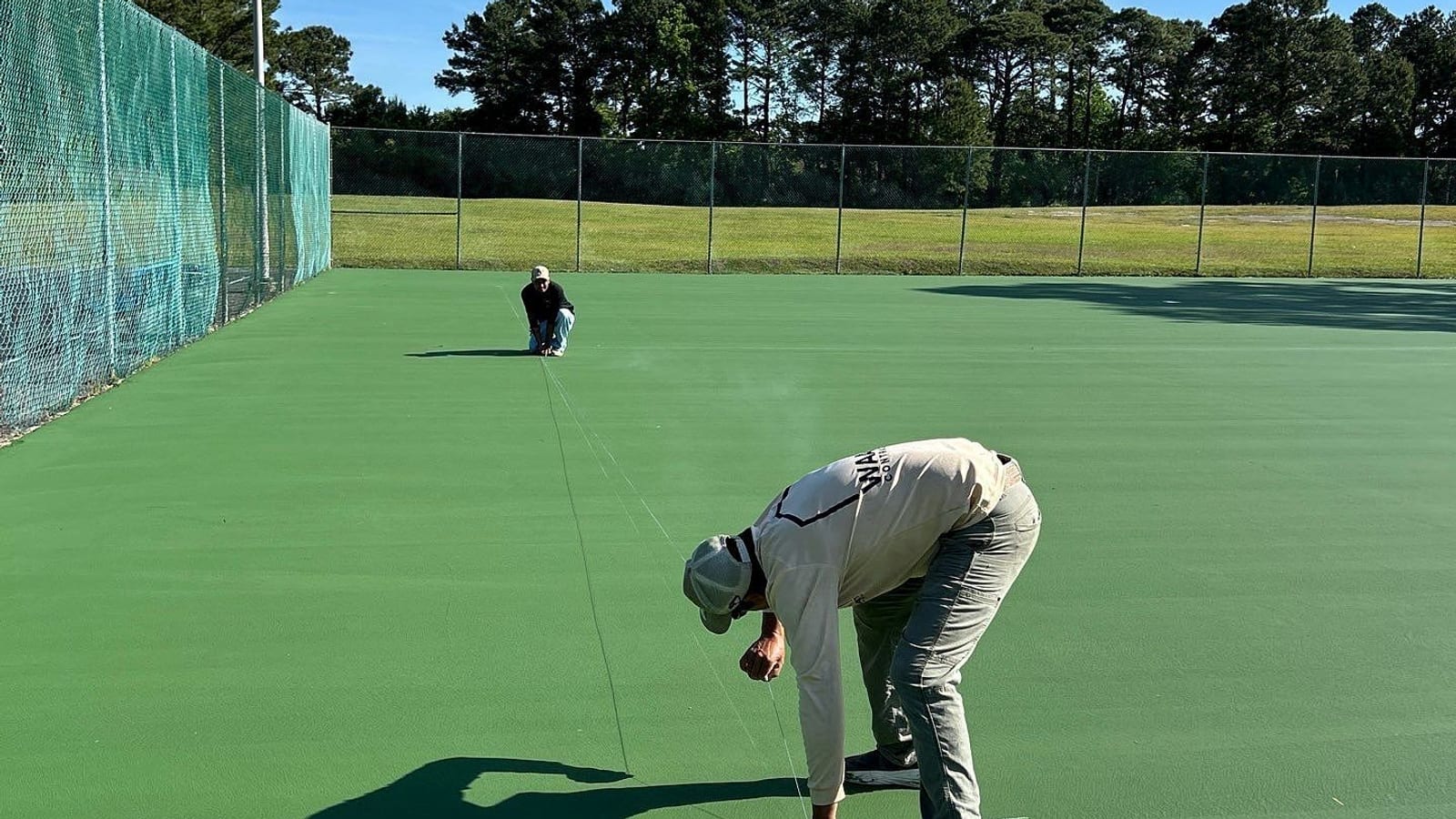 Crew member filling a crack on a green public tennis court before recoating, second crew member at the far fence — Castle Hayne, NC