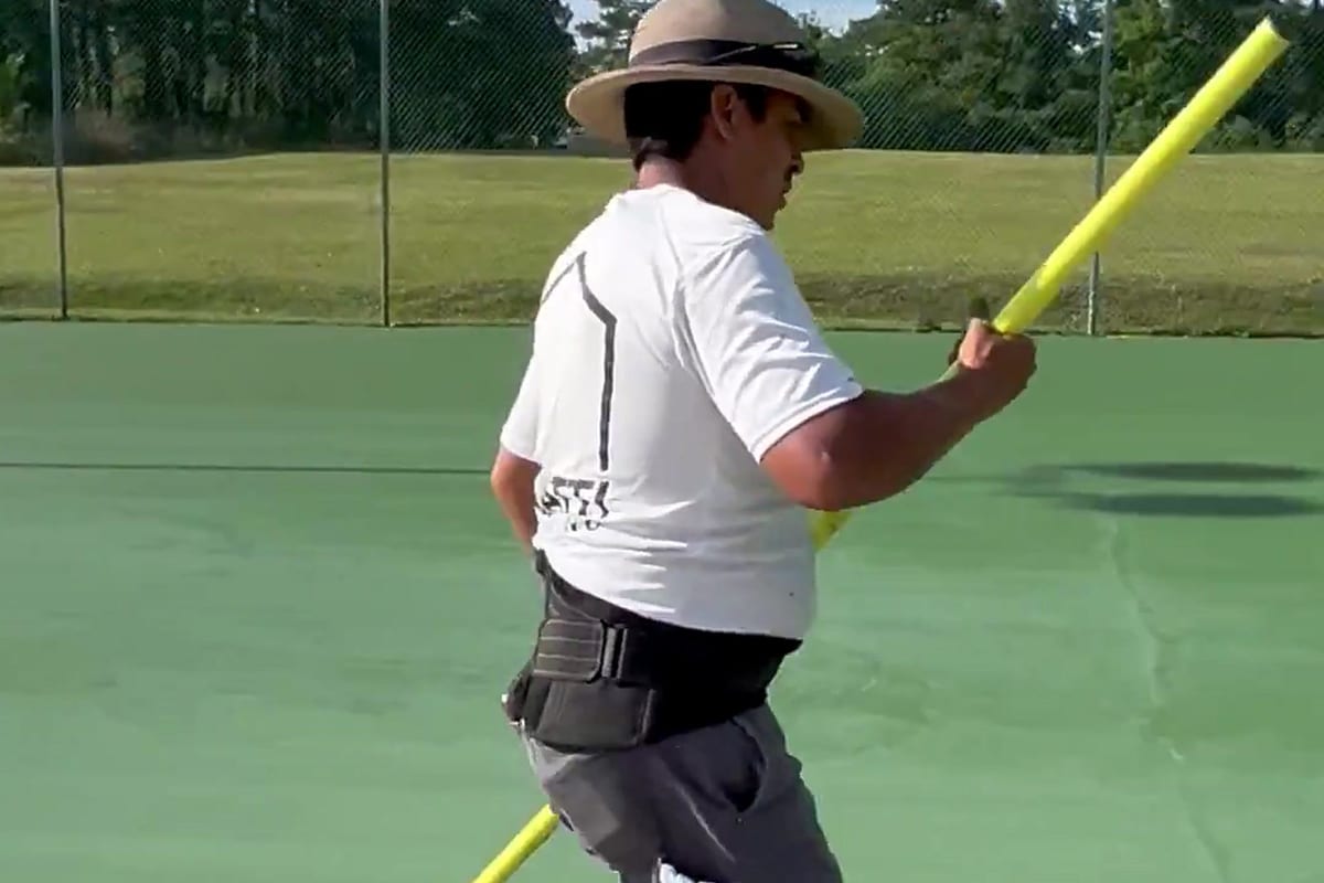 Crew member in a short-sleeve Waatti shirt and sun hat pulling a squeegee across a green tennis court on a summer morning — Castle Hayne, NC
