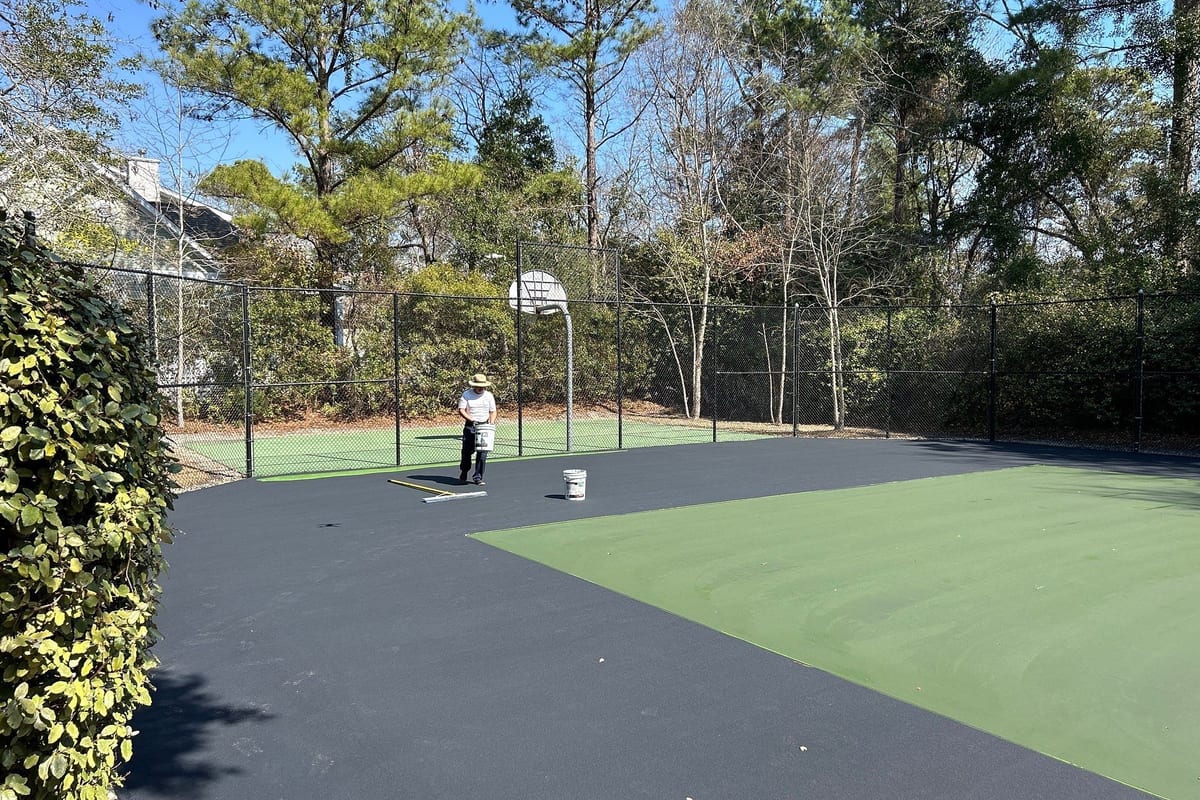 Half-coated tennis court: fresh green acrylic meeting black resurfacer as the crew works toward the far fence — Wilmington, NC