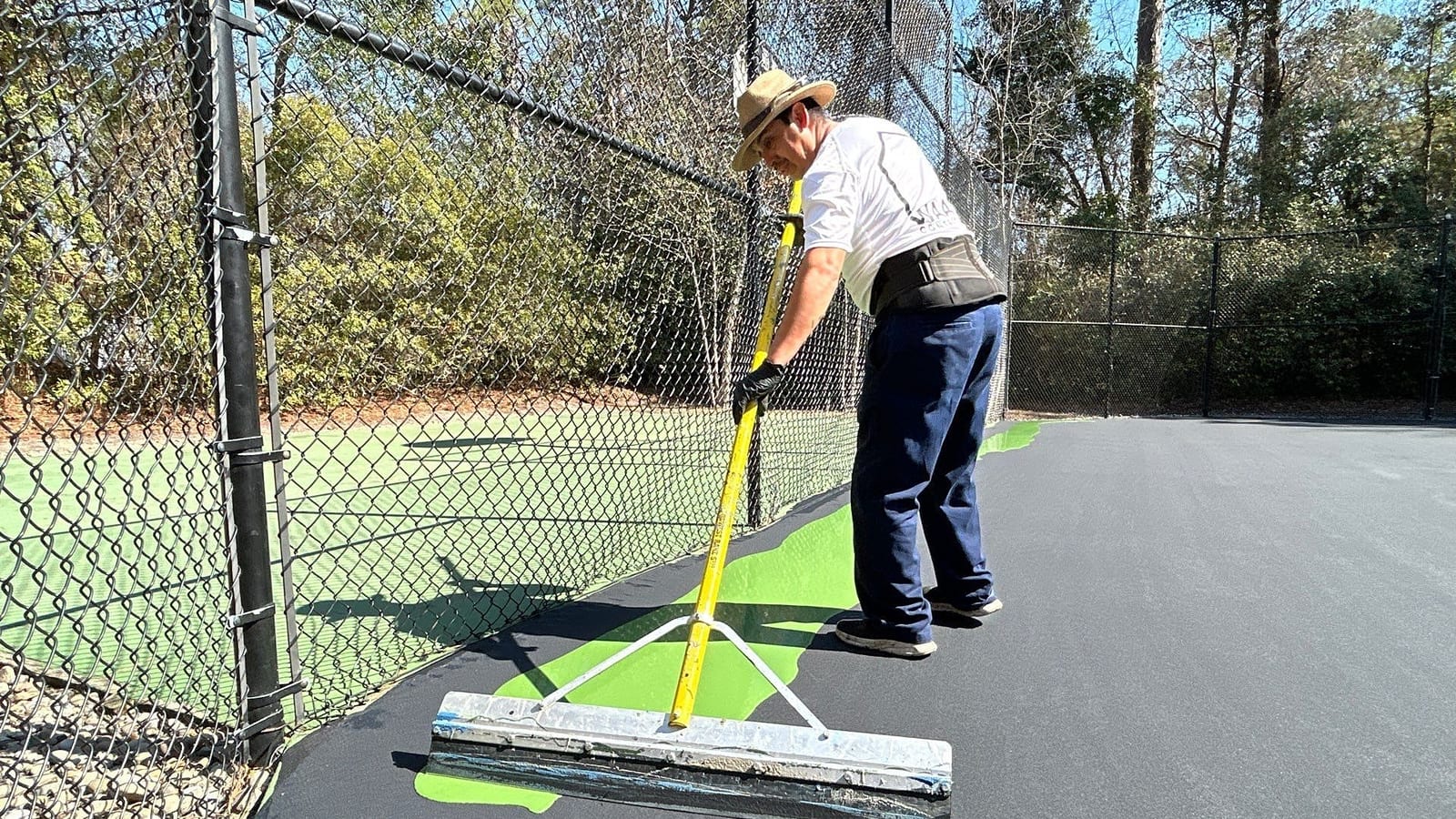 Waatti crew member pulling green acrylic color coat across a tennis court with a squeegee, fence and pines behind — Wilmington, NC