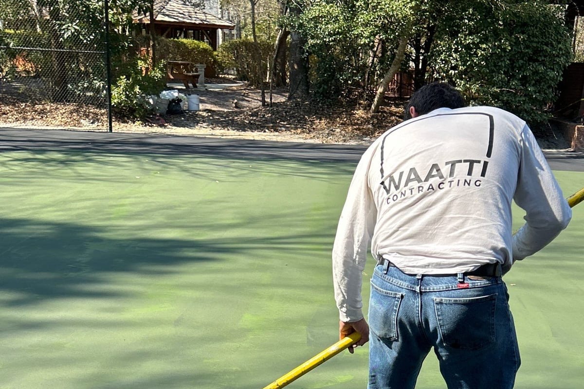Waatti crew member in a white Waatti Contracting shirt pulling bright green acrylic color coating across a court with a squeegee